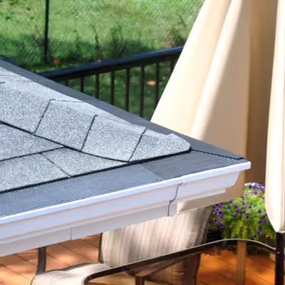 Close-up of a roof corner showing gray asphalt shingles and a black gutter guard installed over a white gutter on a wooden deck.